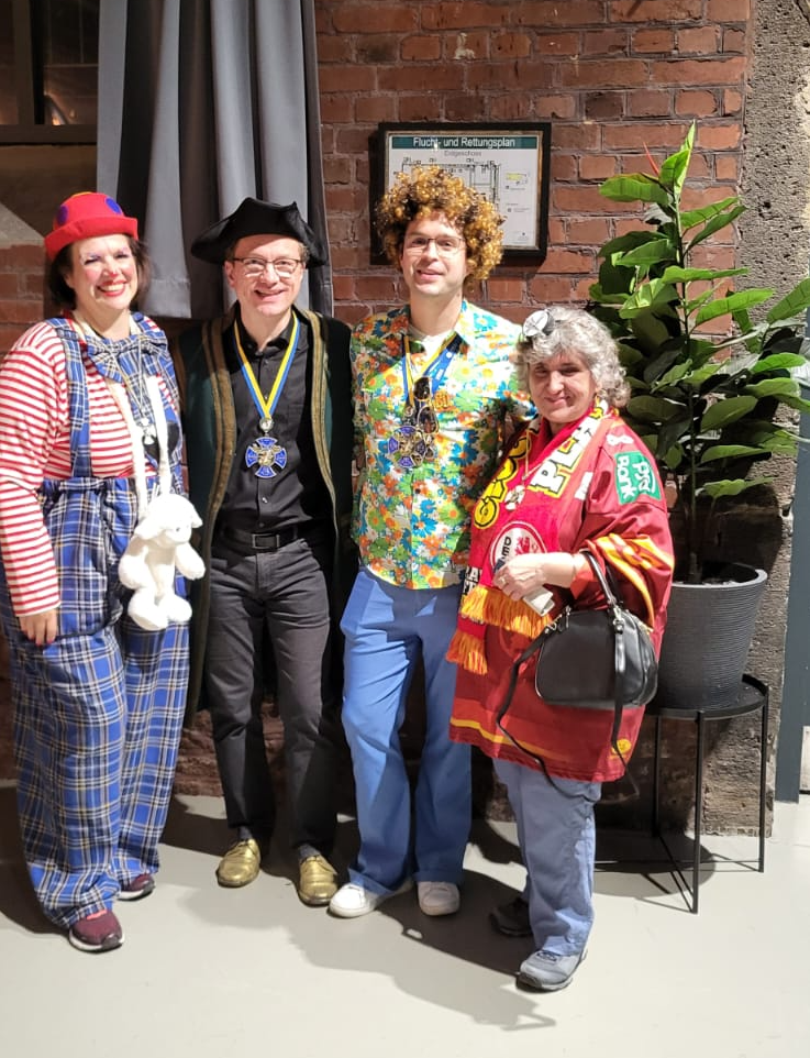 Vier verkleidete Personen stehen nebeneinander vor einer Backsteinwand und lächeln in die Kamera.